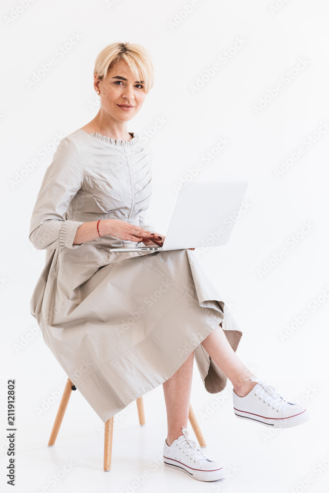 Wall mural Full length portrait of stylish middle aged woman with short blond hair wearing dress sitting in chair and using silver laptop, isolated over white background in studio