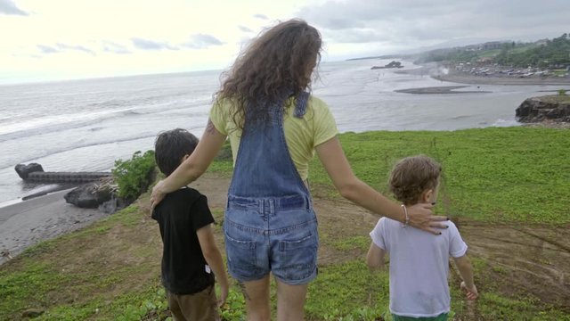 Rear view of young woman in denim romper and two little boys standing on cliff and enjoying view over beach, one of boys pointing at tree