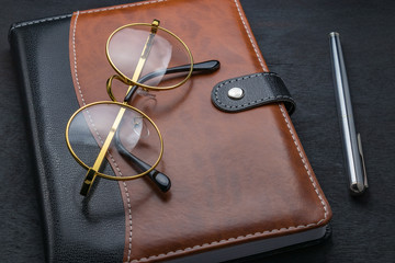 Glasses, notepad, pen lay on a dark table.