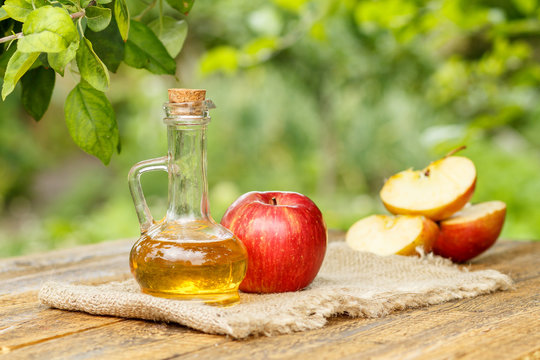 Apple vinegar in glass bottle and fresh red apples on wooden boards in daylight with green natural background
