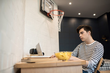 Young male sitting at home at desk with books and bowl of chips and using laptop computer.