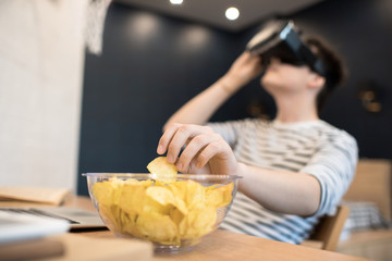 Teenager sitting at home reaching for potato chip in bowl while focused on using virtual reality...