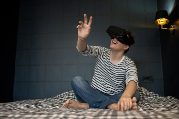Excited teenager extending hand forward while using virtual reality headset sitting on bed in dark...