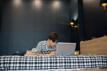 Teenager lying on bed with stack of books using laptop computer and taking notes