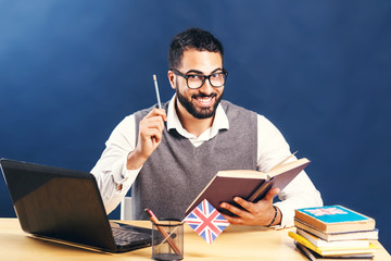 Black-haired man learning english, wearing gray sweater vest and pristine white shirt, smiling at the office desk with laptop before black wall