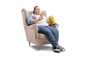 Overweight woman with a drink and a bag of chips sitting in an armchair - obrazy, fototapety, plakaty