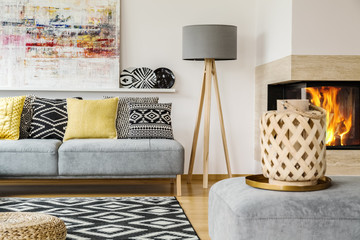 Real photo of a couch with pillows standing next to a lamp and behind a patterned rug and a pouf...