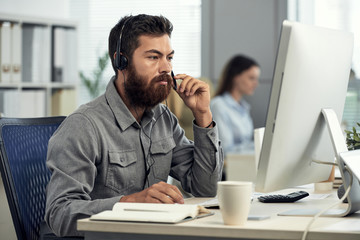 Brutal bearded man in shirt wearing headset and sitting at computer desk in call center 