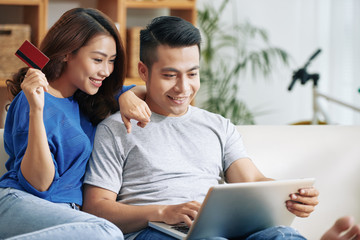 Pretty Asian woman and handsome man relaxing on couch with laptop and surfing Internet while making purchases online