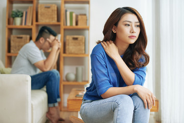Sad Asian woman sitting alone with man on background being in conflict at home