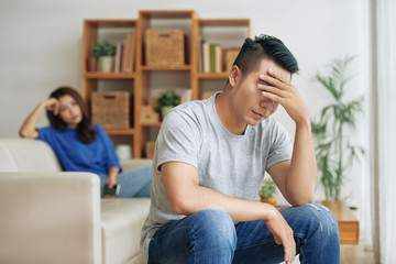 Upset Asian man sitting in living room with woman on background and covering eyes in depression...