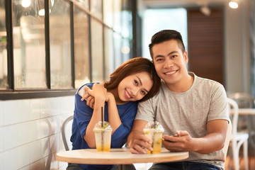 Beautiful young Asian woman and man sitting in cafeteria at table having cool drinks and smiling at camera