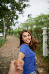 Beautiful Asian woman holding hand of crop anonymous beloved man asking to follow her while strolling in park
