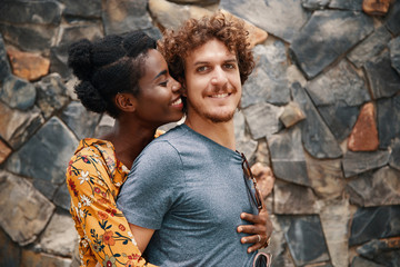 Charming black woman embracing curly man from back and about to kiss while smiling happily outside