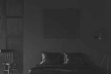 Mockup of black empty poster above bed in dark bedroom interior with chair. Real photo