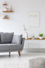 Real photo of a grey couch standing next to  shelf with vases in a white living room with a poster...