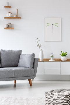 Real photo of a grey couch standing next to  shelf with vases in a white living room with a poster on a wall