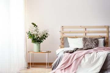 Wooden bedhead with lights by the double bed with fur pillow, grey sheets and pink blanket standing in bright room interior with fresh plants and drapes