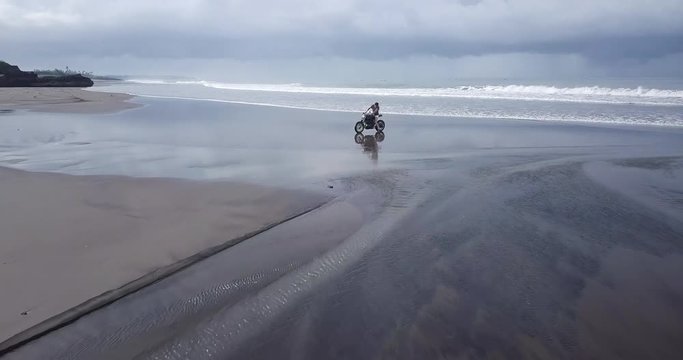 Young handsome hipster man riding modern custom motorcycle cafe racer on the black sand beach near the water. Adventure and travel concept. Surfing spot with ocean waves. Aerial drone footage.