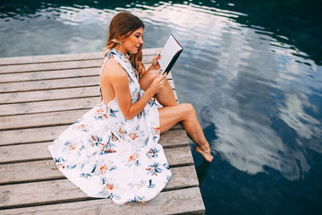 Attractive young woman reading book on dock next to the lake