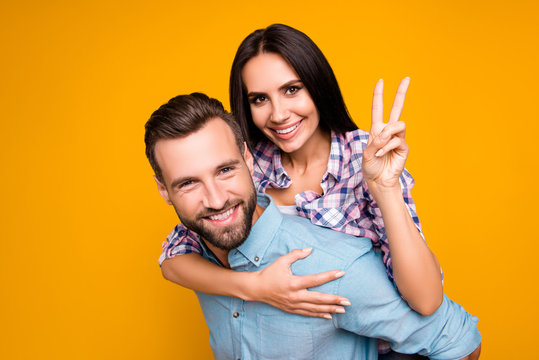 Portrait of friendly lovely couple gesturing v-sign with two fingers, handsome man carrying on back attractive woman isolated on vivid yellow background