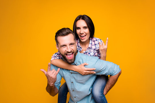 Portrait of funky crazy couple gesturing rock and roll sign yelling, handsome man carrying on back pretty woman looking at camera isolated on vivid yellow background