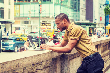 Young African American Businessman with beard traveling, working in New York, wearing green short sleeve shirt, black pants, bending back over half wall fence in Manhattan, working on laptop computer