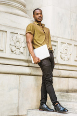 Young African American Male College Student with beard studying in New York, wearing green short sleeve shirt, black pants, leather shoes, carrying laptop computer, standing against white marble wall