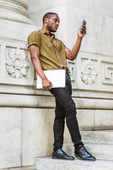 Young African American Man studying in New York, wearing green short sleeve shirt, black pants, leather shoes, carrying laptop computer, standing against white marble wall, texting on cell phone..