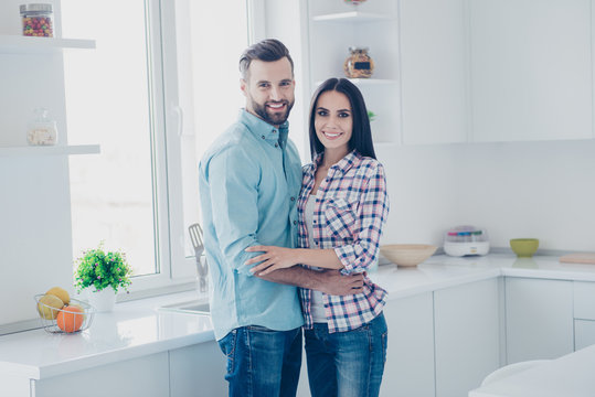 Portrait of attractive beautiful couple with modern hairstyle in casual clothes hugging looking at camera standing in modern white kitchen with interior