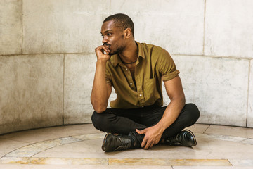 Young African American Man wearing green short sleeve shirt, black pants, leather shoes. legs crossed, sitting on marble ground in New York, raising arm, hand touching mouth and chin, thinking..