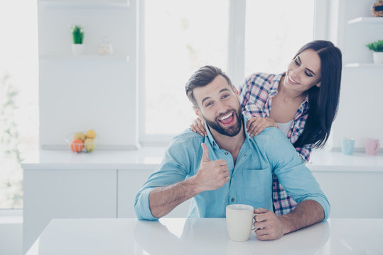 Portrait of excited cheerful man sitting in modern white kitchen drinking tea gesturing thumb up symbol enjoying care of woman. Domicile togetherness friendship concept