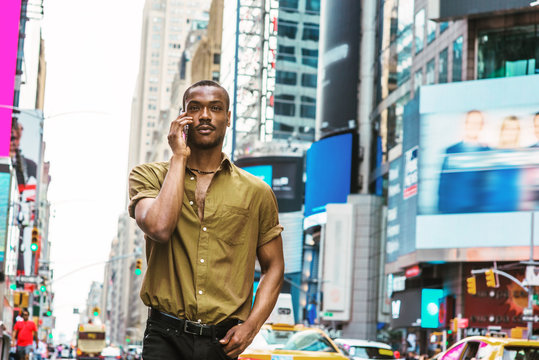 Young African American Man traveling in New York, wearing green short sleeve shirt, walking on busy street in Times Square of Manhattan, talking on cell phone. High buildings, cars on background..