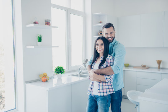 Portrait of lovely modern couple embracing in white kitchen with interior looking at camera wearing casual clothes enjoying time together