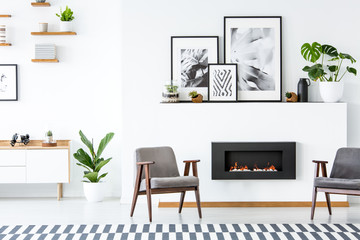 Black fireplace between grey armchairs in apartment interior with posters and plants. Real photo