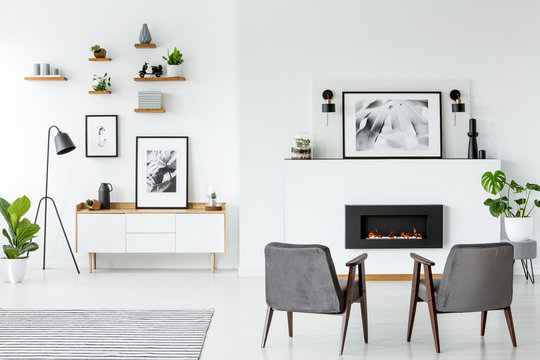 Grey armchairs in front of fireplace under poster in white apartment interior. Real photo