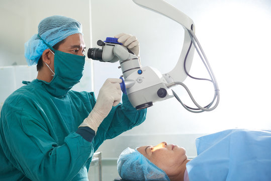 Focused Asian man in mask and coat of surgeon looking at eyes of woman on table using operating microscope 