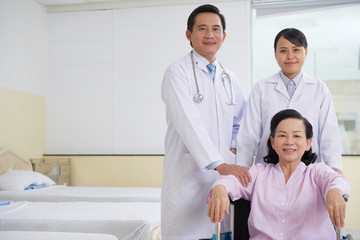 Obraz premium Cheerful Asian man and woman in medical gowns standing with mature woman in wheelchair in hospital ward smiling at camera