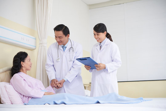 Asian Smiling Doctors Visiting Mature Woman In Hospital Ward And Taking Care Of Health 