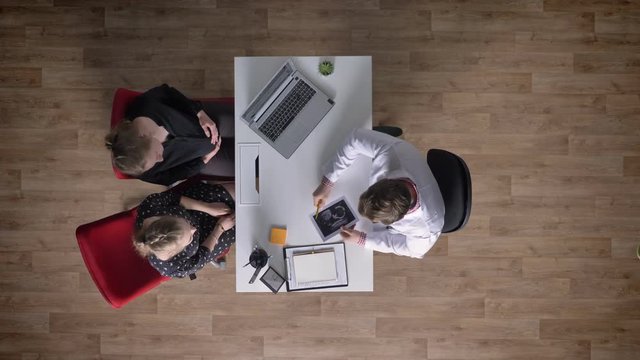 Young pregnant woman on consultation with doctor, two women sitting at table and speaking with doctor, topshot, happy