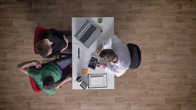 Young doctor advising couple of man and pregnant woman, topshot, happy, hugging, medical concept