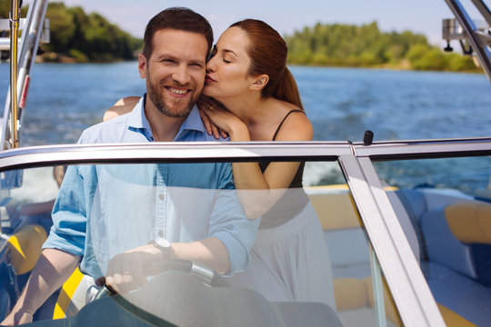 Love you. Affectionate young woman giving her husband a kiss on the cheek while he sailing a boat and smiling