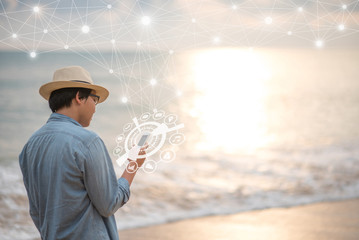 Young Asian man using smartphone with glowing mobile application icon hologram on tropical beach during sunset. Global communication network and Internet of things (IOT). Summer vacation lifestyle