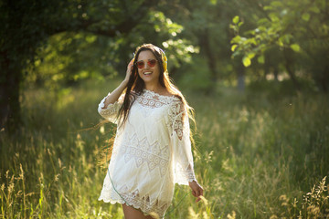 Pretty girl dances with headphones on outdoors in nature. Beautiful young woman wearing white dress and sunglasses dancing in forest glade with headphones on. Sun rays glooming through trees.