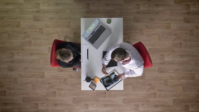 Doctor in white coat showing x-ray scan to young woman, sitting at table in office, topshot, medical concept