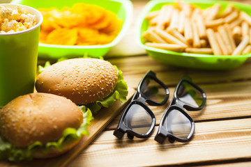 Weekend at home, leisure lifestyle, TV,  fast food concept. Set of 3D glasses, remote controllers and snacks in green bowls: chips, popcorn, burgers on the wooden background. Top view. Close up