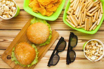 Weekend at home, leisure lifestyle, TV,  fast food concept. Set of 3D glasses, remote controllers and snacks in green bowls: chips, popcorn, burgers on the wooden background. Top view. Close up