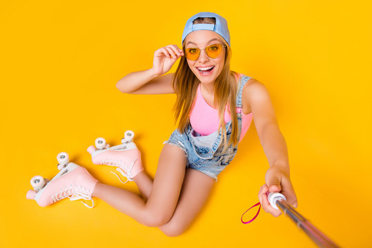 Self portrait of toothy joyful girl shooting selfie on front camera sitting on floor ground wearing roller skates isolated on yellow background, photography concept