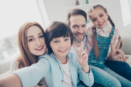 Self portrait of friendly lovely family with two kids hugging sitting on couch shooting selfie on front camera having video-call gesturing v-sign hi symbol. Little blogger cool son concept