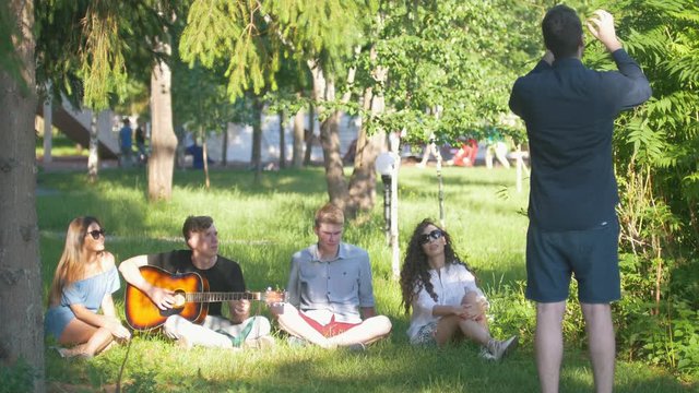 Young man shows pantomime in front of friends sitting on the grass in summer park